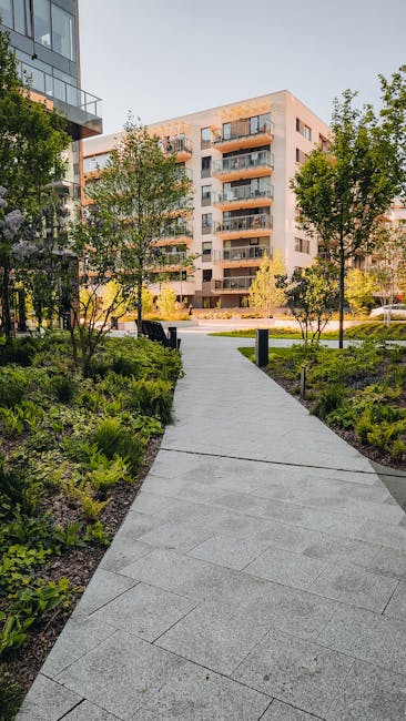 A well-maintained urban park in Barking Riverside estates featuring a lush, green lawn bordered by mature trees with budding leaves, indicative of springtime. The scene includes a paved winding pathwa